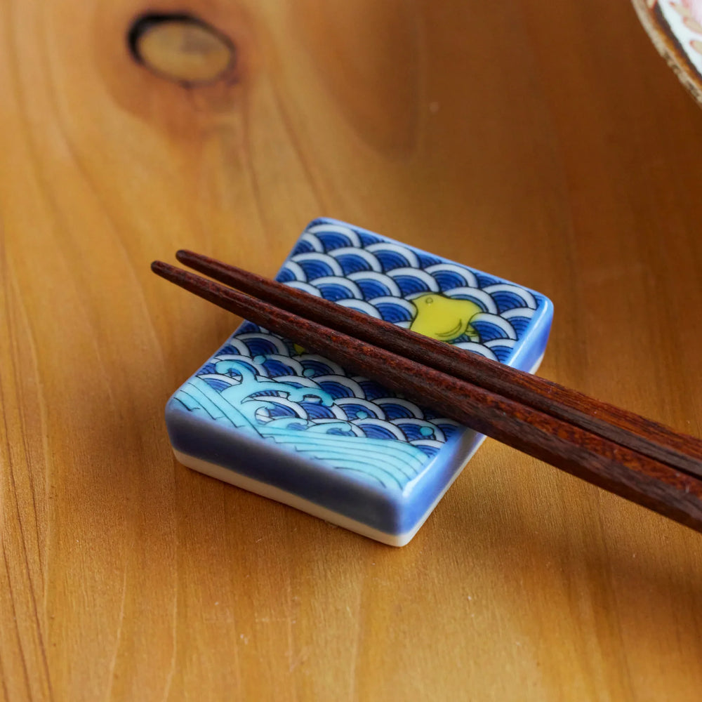 Porcelain chopstick rest with blue wave patterns and a yellow shorebird, holding wooden chopsticks on a wooden table.