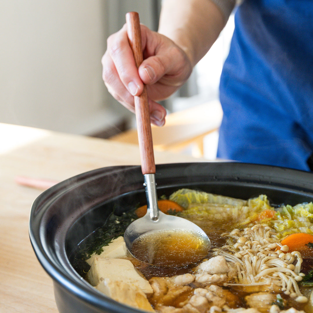 Stainless steel soup ladle with wooden handle scooping broth from Japanese donabe hot pot