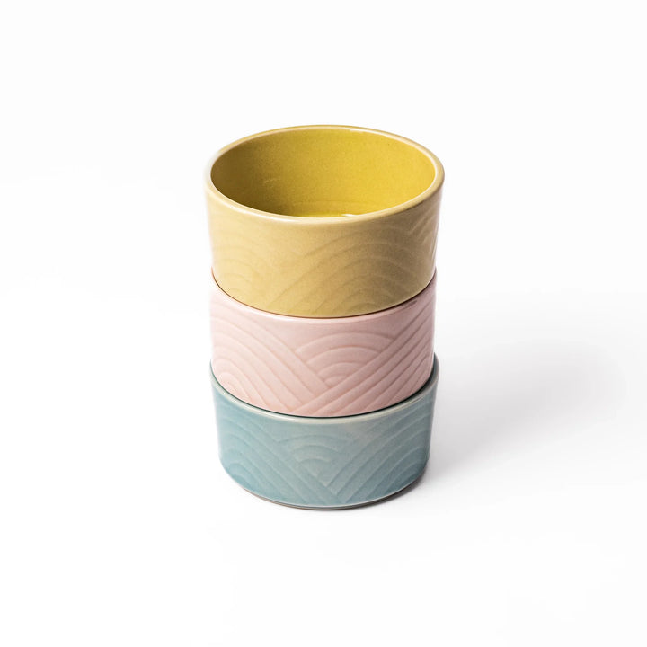 Three stackable medium porcelain bowls in pastel yellow, pink, and blue, featuring embossed Seigaiha wave patterns and a smooth matte finish.