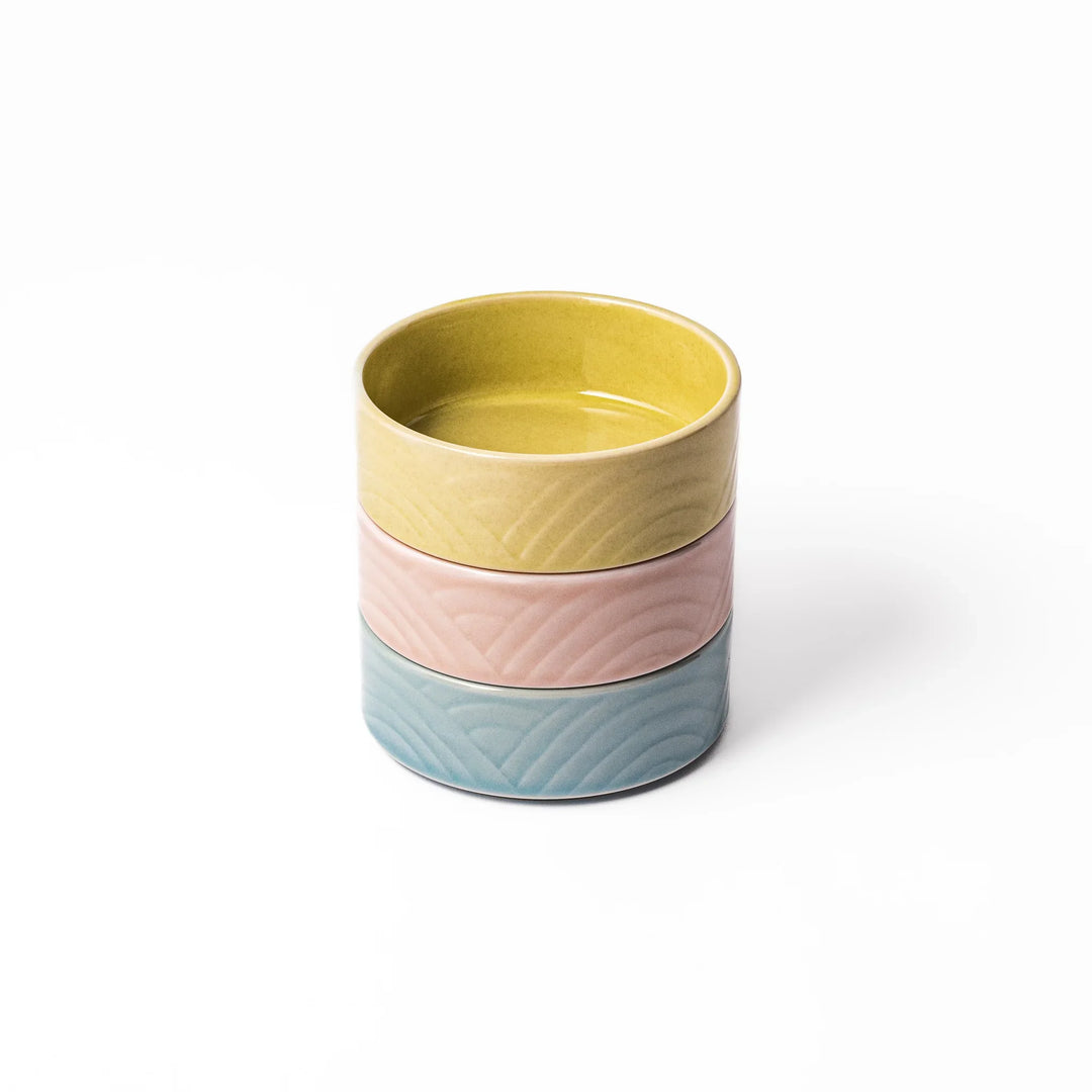 Three small porcelain bowls in yellow, pink, and blue stacked neatly, each featuring an embossed Seigaiha wave pattern for a soft, modern look.
