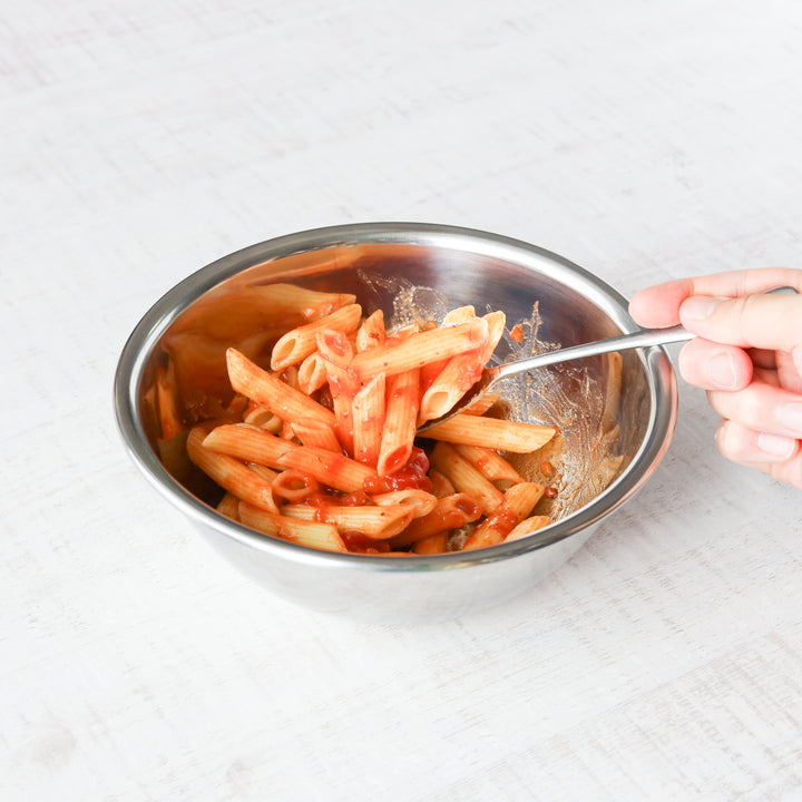 Stainless steel mixing bowl being used to toss penne pasta with tomato sauce, highlighting practical kitchen prep use.