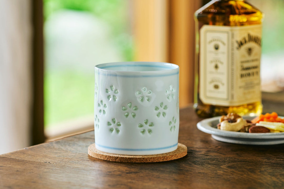 Light blue ceramic cup with delicate cutout sakura pattern on a cork coaster beside a bottle and snack plate on a wood table