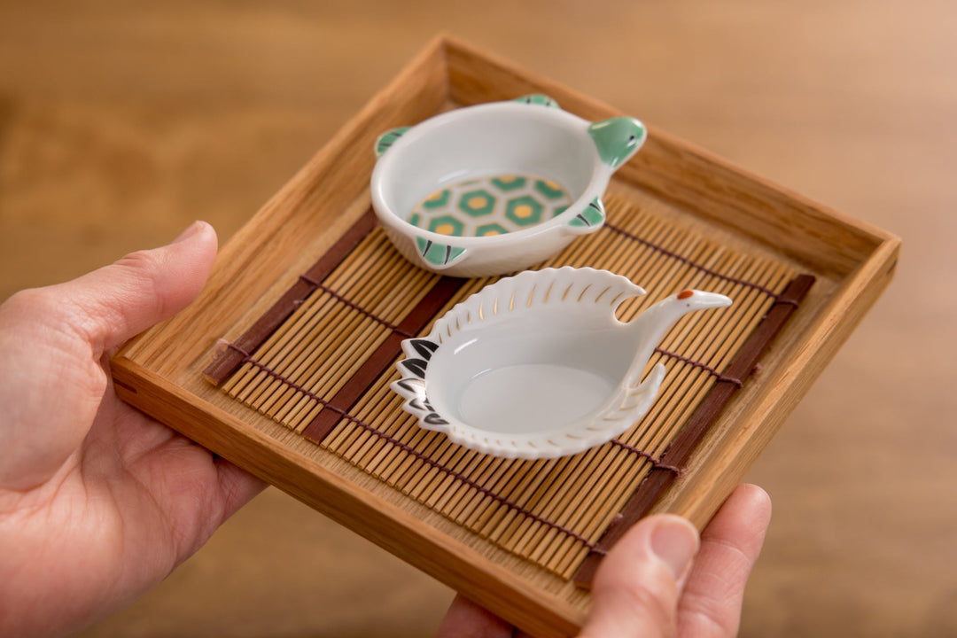 Hands holding a bamboo tray with two small Japanese ceramic dishes shaped like a turtle and a crane.