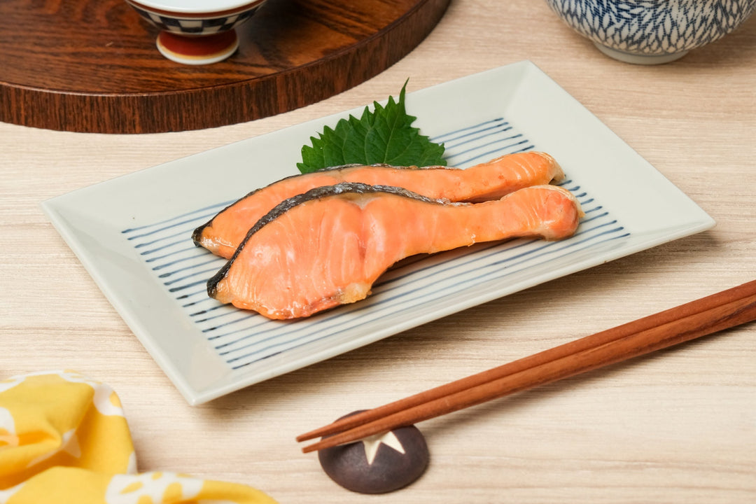 Rectangular plate with grilled salmon and shiso leaf with chopsticks beside small bowls and rice on a light table setting