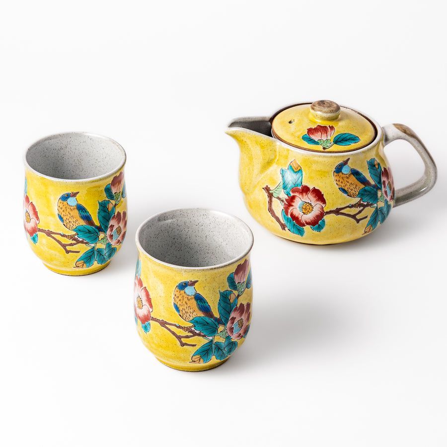 Three piece ceramic tea set with a warm yellow glaze featuring hand-painted camellia flowers and birds on a teapot and two matching cups.