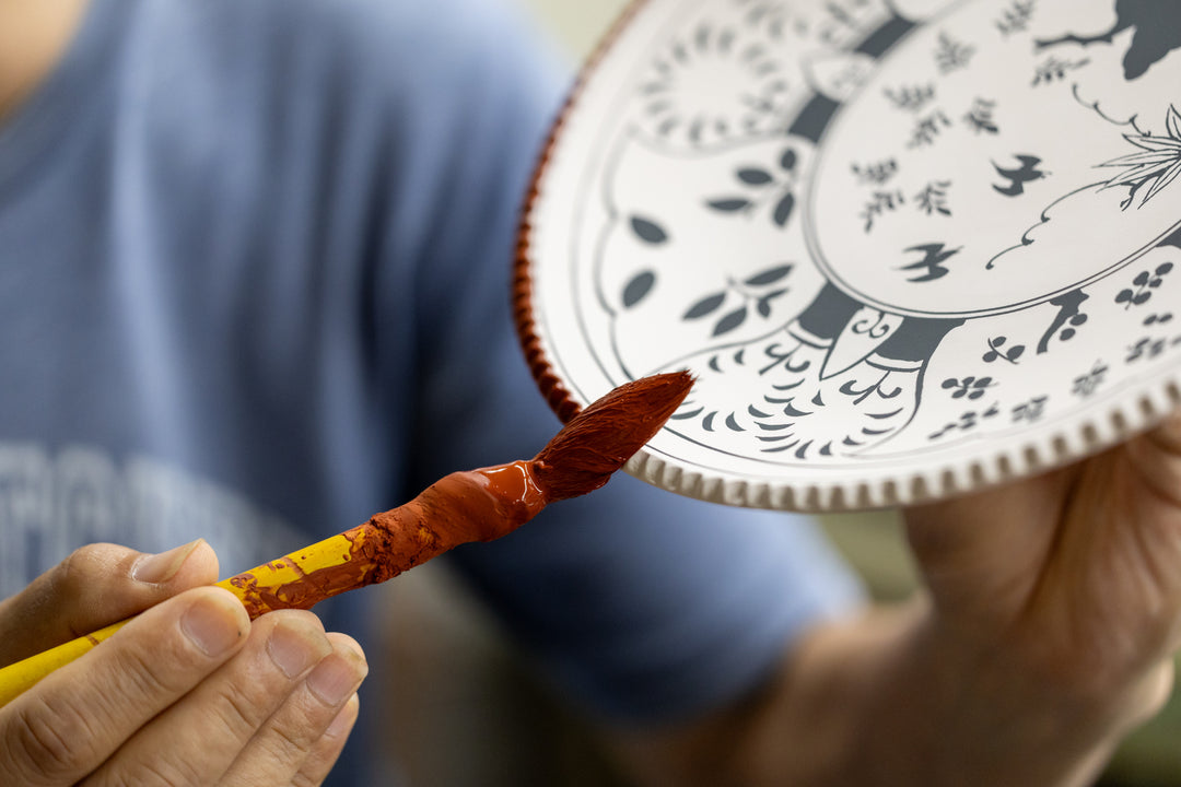 Artisan hand-painting a ceramic plate with red pigment, adding detailed patterns to the traditional Japanese design.