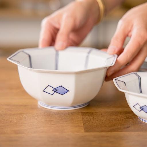 A person holds an octagonal cereal bowl decorated with blue diamonds, alongside another similar bowl on a wooden surface, showcasing its elegant design and shape.