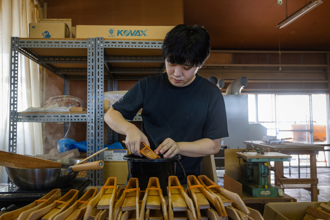 Artisan crafting hinoki wood utensils, dipping pieces into lacquer in a workshop filled with tools and steam.