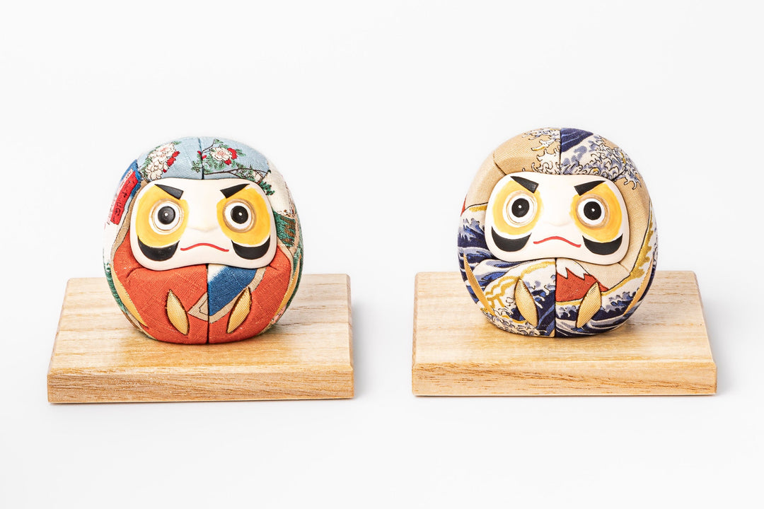 Two traditional Japanese fabric daruma dolls with colorful patterns displayed on wooden stands side by side