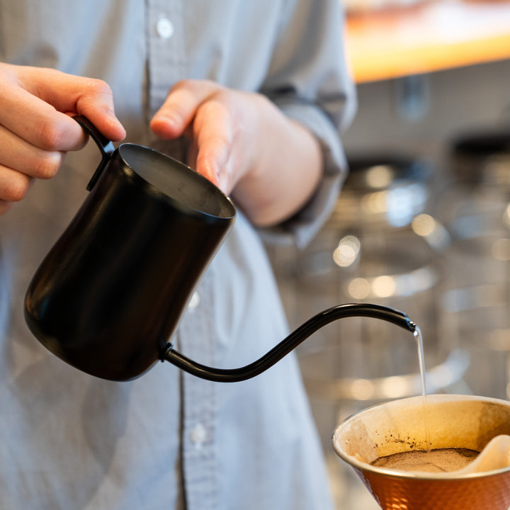 Person brewing coffee with a Japanese matte black precision pour kettle, pouring hot water into a dripper.