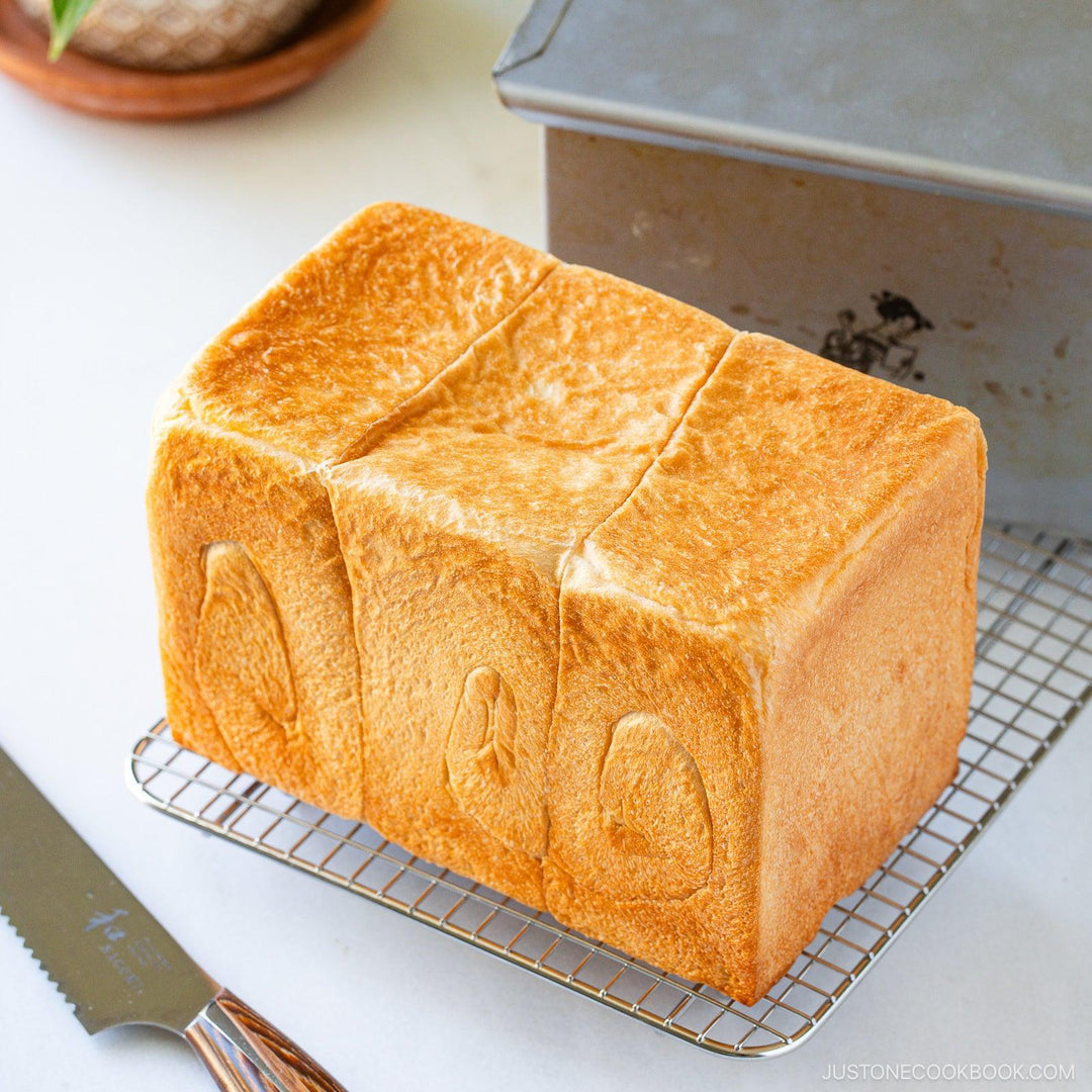 Japanese shokupan loaf pan with baked milk bread on cooling rack with bread knife and metal bread box