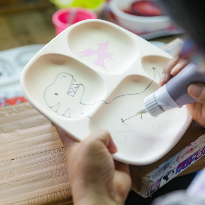 Artisan adding fine painted lines to a dinosaur illustration on a kids section plate in the workshop