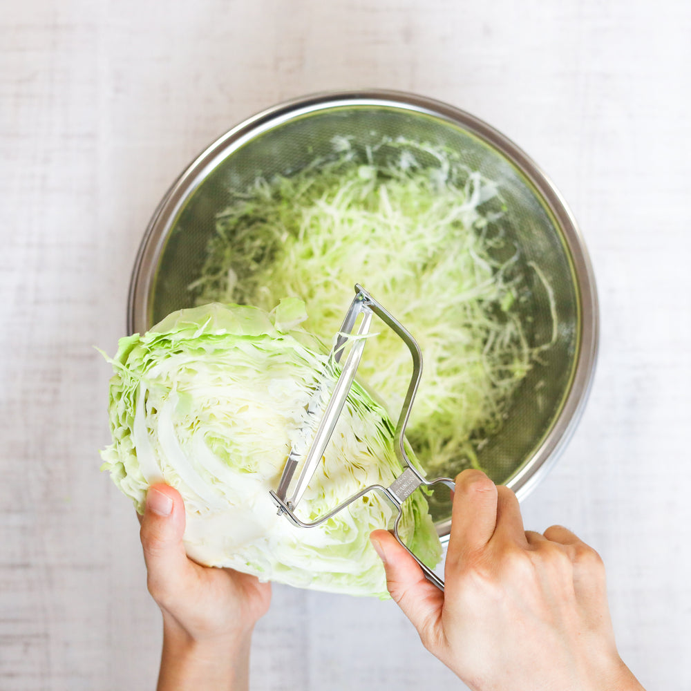 Person using a vegetable peeler to shred green cabbage into a metal bowl.