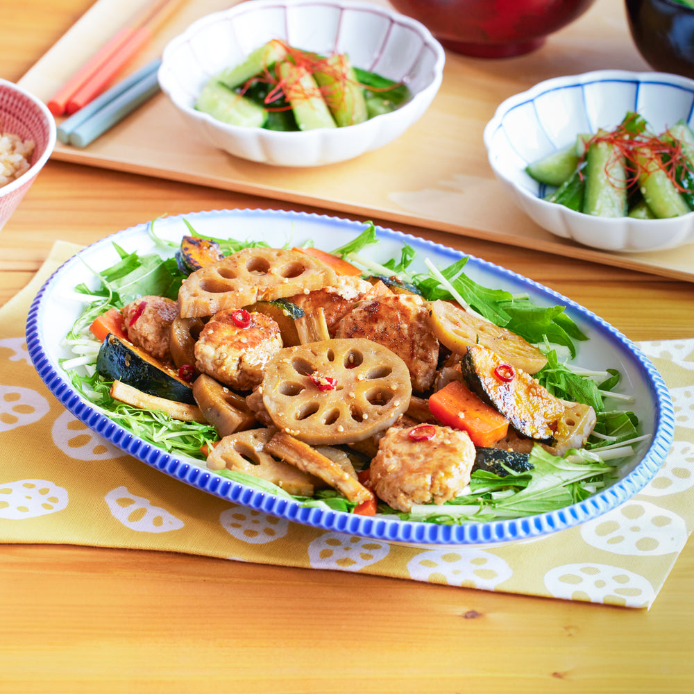 A blue-rimmed Japanese oval plate filled with stir-fried lotus root, chicken meatballs, and colorful vegetables.