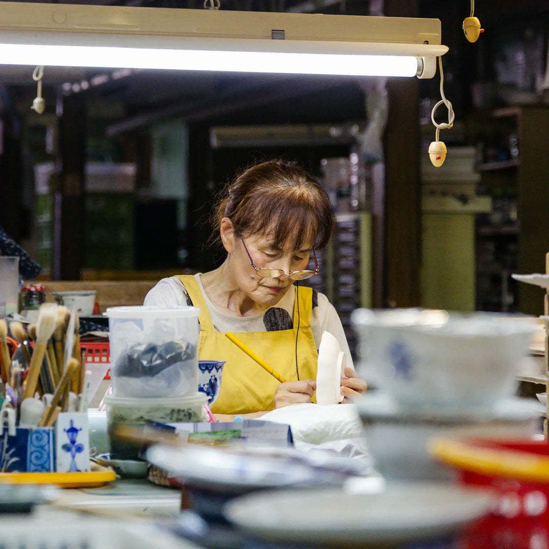 Artisan hand painting porcelain tableware at a Japanese kiln workshop surrounded by brushes and tools.