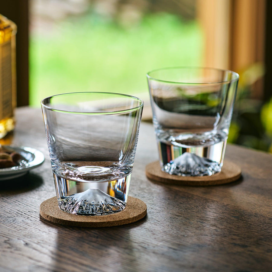 Two clear Japanese whiskey glasses with Mt. Fuji-shaped bases, set on cork coasters on a wooden table.