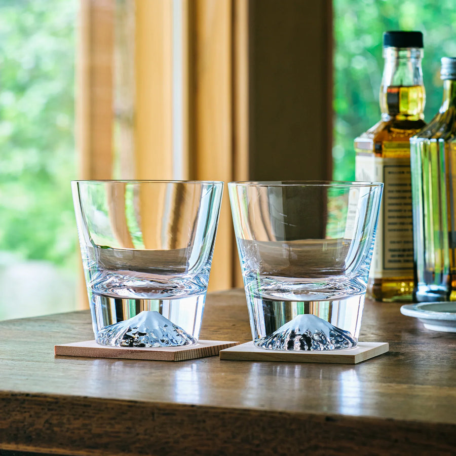 Japanese whiskey glasses with Mt. Fuji design on wooden coasters, placed on a table by the window.