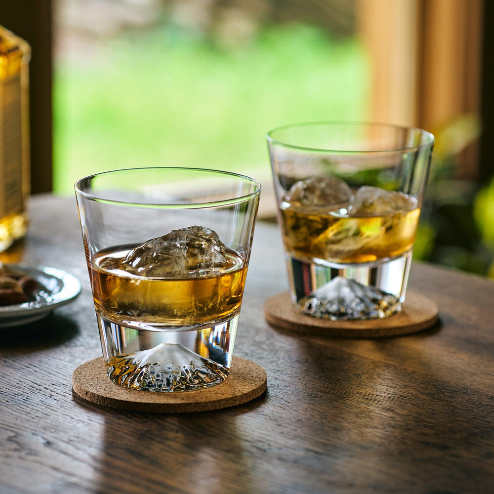 Two Japanese whiskey glasses with Mt. Fuji-shaped bases, filled with whiskey and ice, on cork coasters.