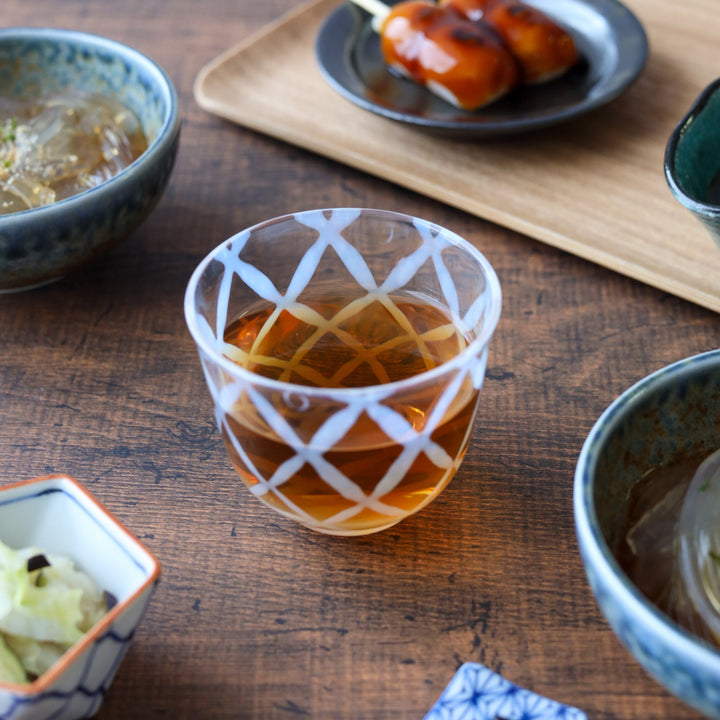 Clear glass tea cup with a vintage-style white lattice pattern, filled with amber tea and set among traditional Japanese dishes on a wooden table.
