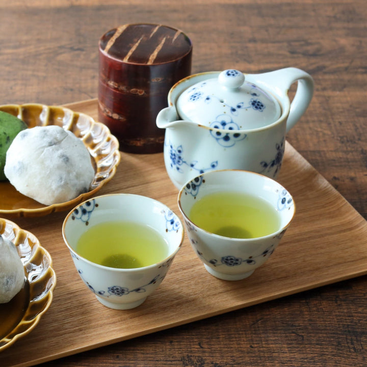 Wooden tray with a floral teapot, matching cups filled with green tea, dessert plates holding mochi, and a white bud vase with delicate flowers.