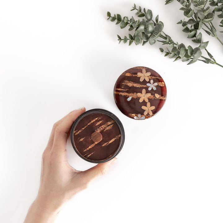 Hand holding a cherry bark tea caddy with a lid adorned with mother-of-pearl cherry blossoms, a beautiful and functional piece for storing precious tea leaves.