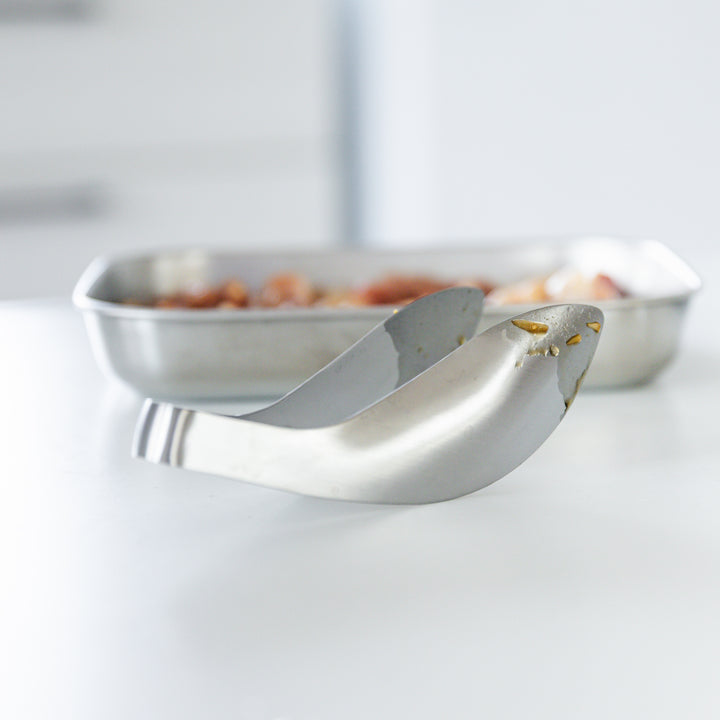 A pair of stainless steel tongs resting on a white surface, with a stainless tray of marinated ingredients blurred in the background.