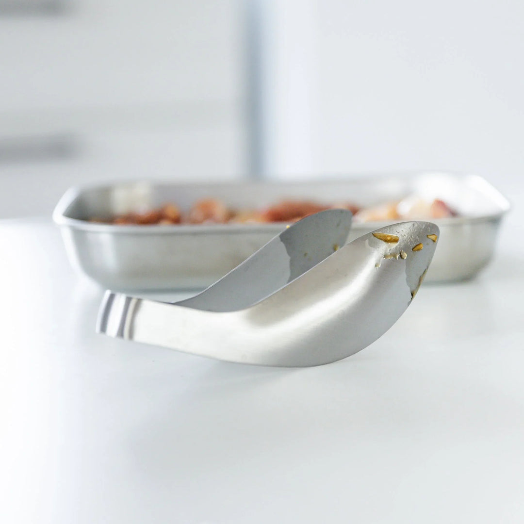 Japanese stainless steel kitchen tongs with curved tip design for easy grip and precise cooking, placed beside a metal tray