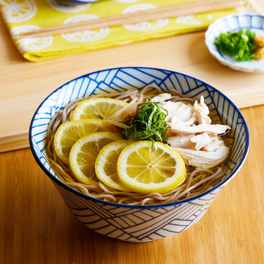 Blue and white ramen bowl with a bold basket weave pattern, filled with soba, lemon slices, shredded chicken, and herbs on a wooden table.