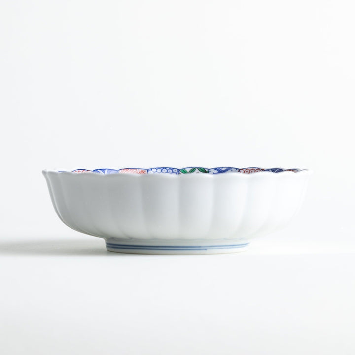 A round, deep cereal bowl with a scalloped edge, featuring a central floral design surrounded by a colorful pattern in blue, red, and green on a white background.