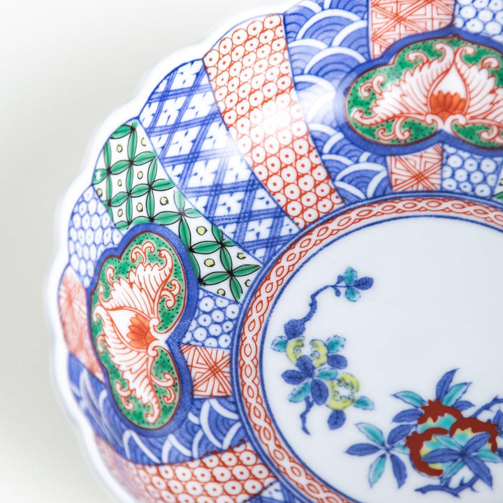 A round, deep cereal bowl with a scalloped edge, featuring a central floral design surrounded by a colorful pattern in blue, red, and green on a white background.