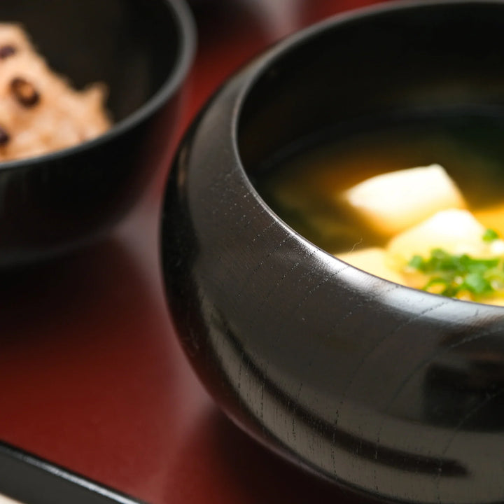 Black lacquered oryoki bowl filled with tofu miso soup showcasing fine wood grain and glossy handcrafted finish