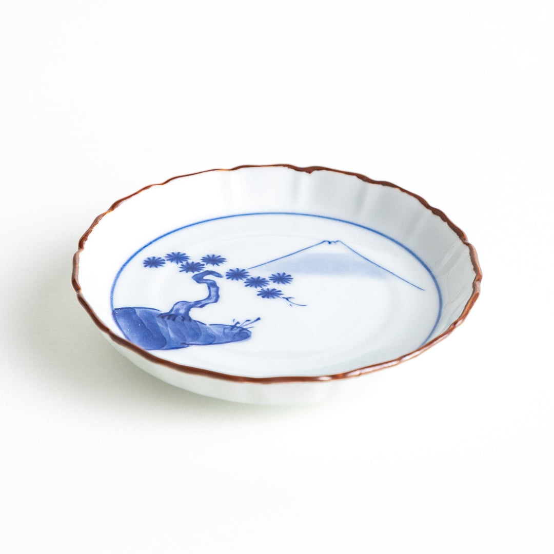 Angled view of a porcelain sauce dish displaying its slightly raised rim and blue Mount Fuji and pine tree motif, with a brown accent on the edge.