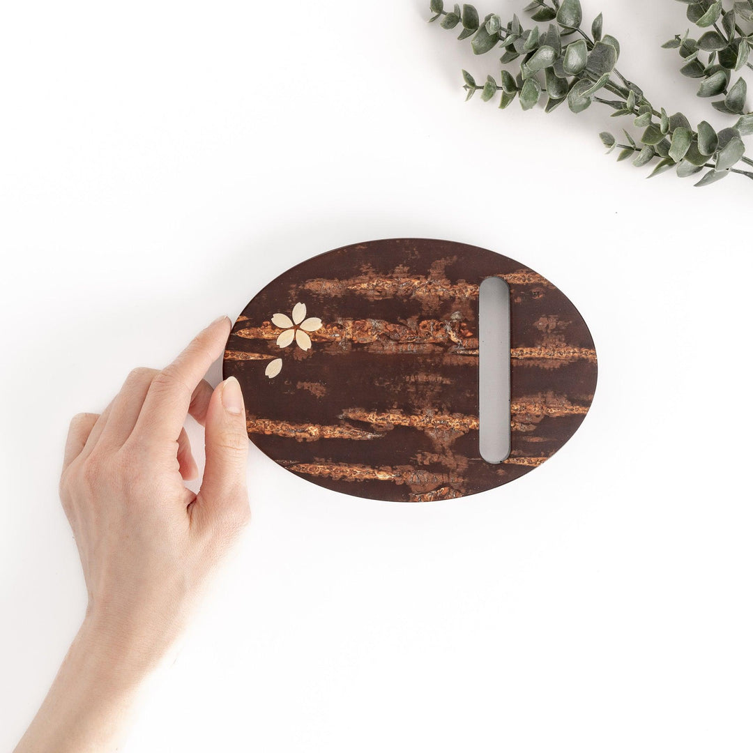 Hand touching an oval-shaped cherry bark teapot rest with a delicate cherry blossom inlay, perfect for protecting surfaces from heat and adding a touch of nature to your tea setting.