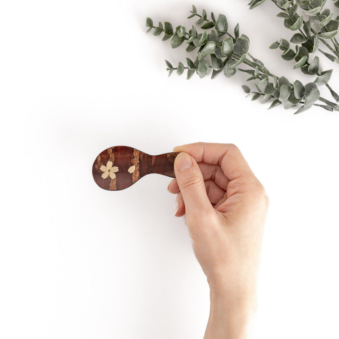 Hand holding a handcrafted cherry bark tea scoop with a delicate cherry blossom inlay, perfect for measuring loose leaf tea for a perfect cup.