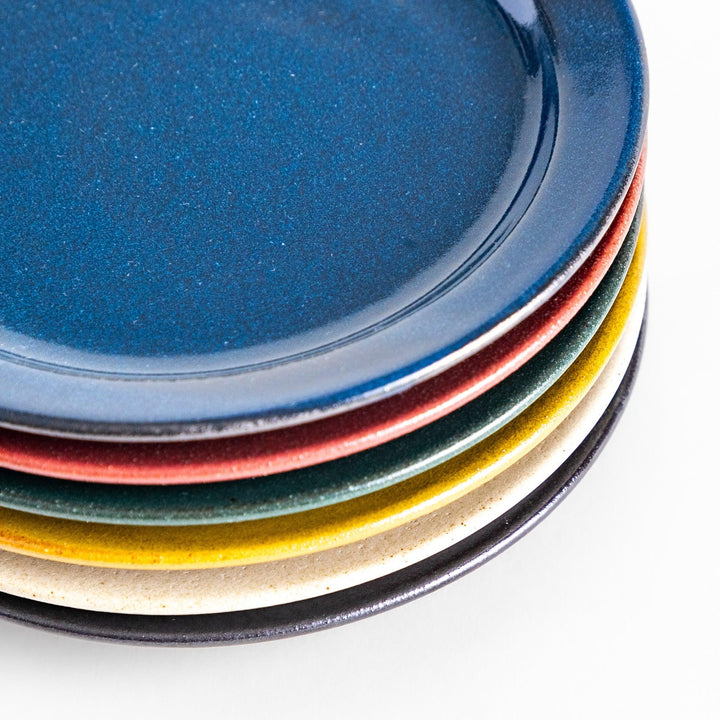 A close-up view of six colorful dessert plates in blue, red, green, yellow, white, and black, each featuring a smooth, glossy finish, stacked neatly to showcase their vibrant hues.