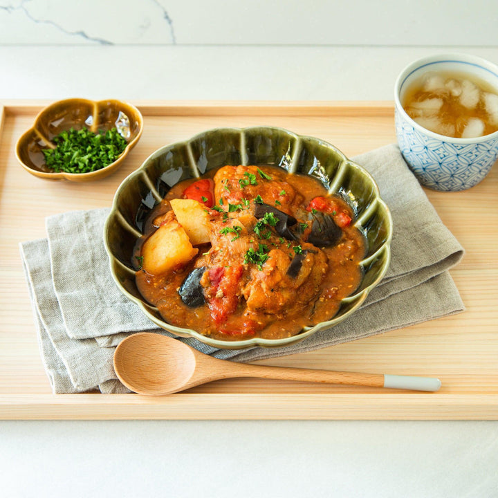 Traditional blue-patterned tea bowl filled with iced tea, placed on a wooden tray alongside a green pasta bowl of miso tomato chicken stew and a small dish of chopped herbs.