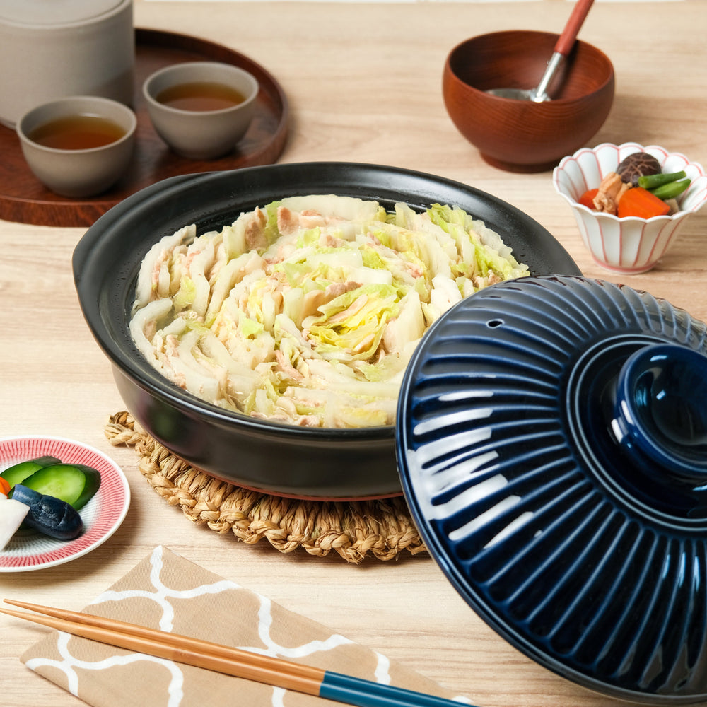Navy Japanese clay pot filled with layered napa cabbage and pork hot pot, placed on a straw trivet with tea, pickles, and side dishes for cozy dining.