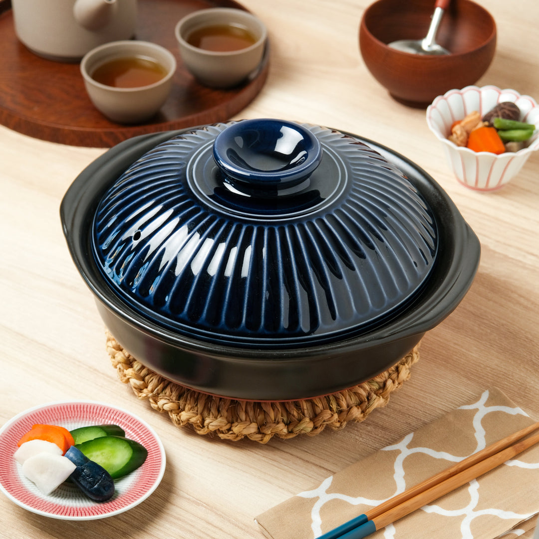 Ginpo Kikka Family-Size Donabe in navy with a ribbed lid, set on a straw trivet alongside pickles, simmered vegetables, tea, and chopsticks for authentic Japanese dining.