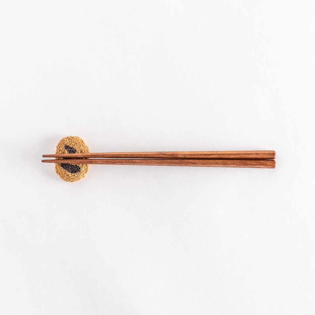 A wider-angle view of a Japanese croquette-shaped chopstick rest with chopsticks placed on top, showing the textured breadcrumb surface and black swirl design.