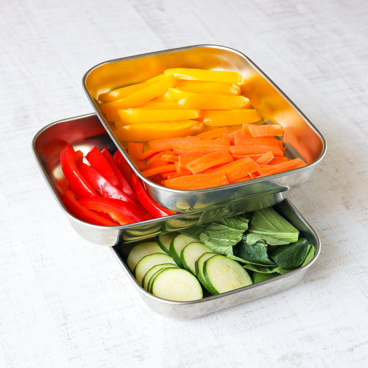 Colorful sliced vegetables—red and yellow bell peppers, carrots, zucchini, and greens—neatly arranged in stackable stainless steel prep trays.