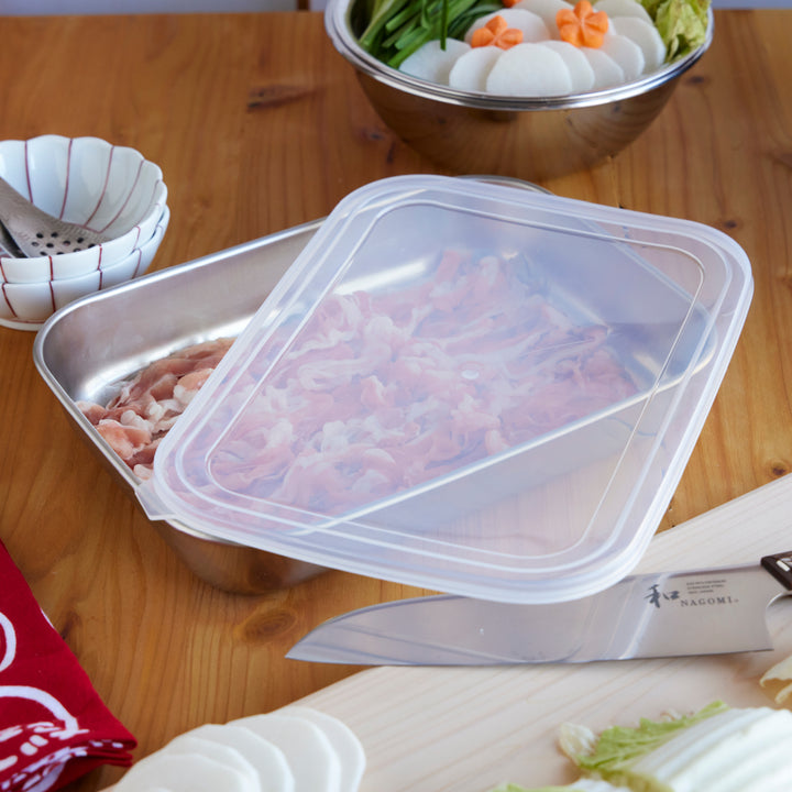 Stainless steel tray with lid holding sliced pork for hot pot, shown with Japanese Nagomi knife, vegetables, and sauce bowls.