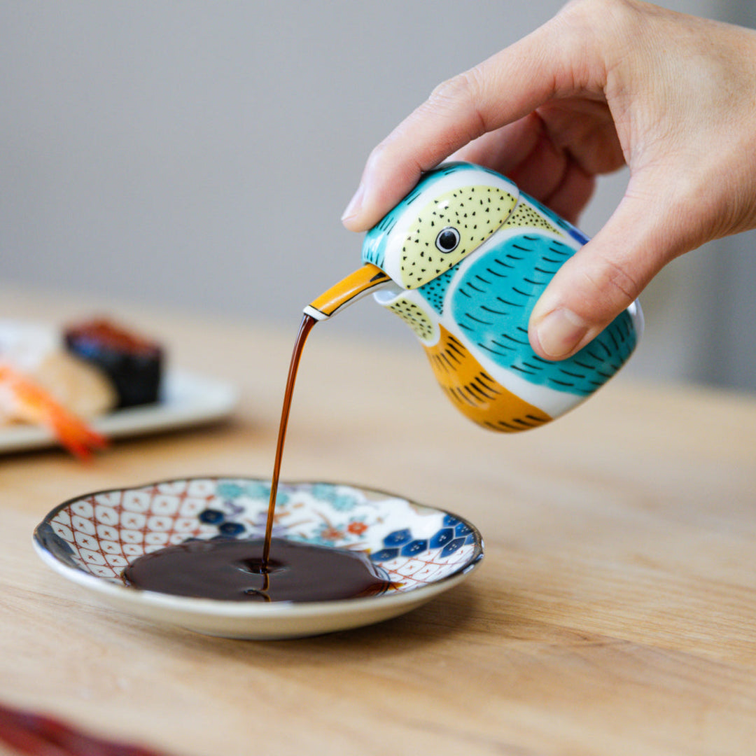 Pouring soy sauce from a blue bird ceramic kingfisher dispenser into a small dish next to fresh sushi