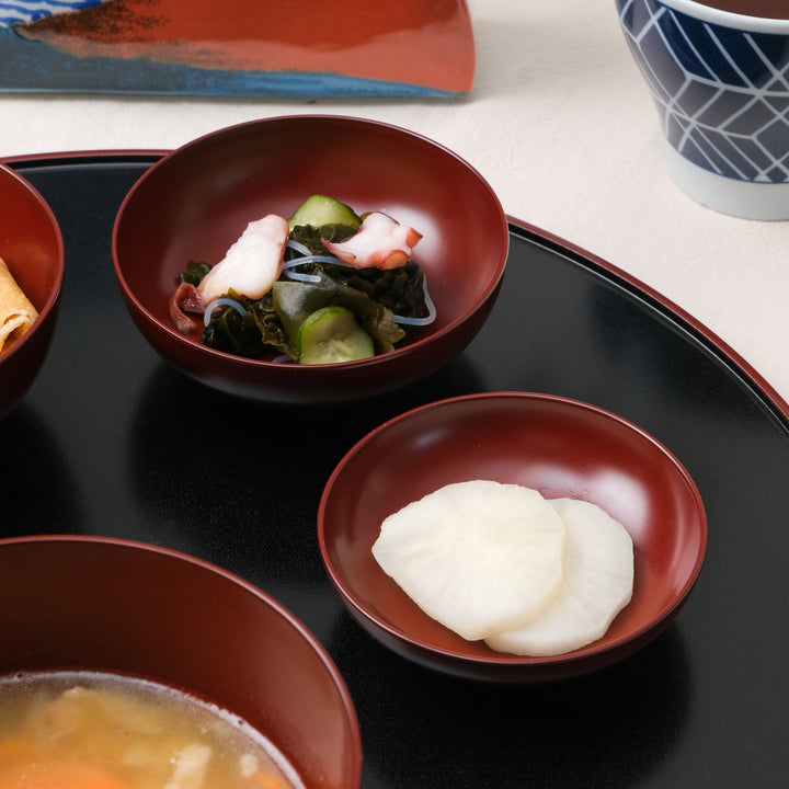 Close-up of Japanese Oryoki side dishes featuring sunomono with octopus, cucumber, and seaweed, and crisp daikon pickles served in red lacquer bowls on a black tray.