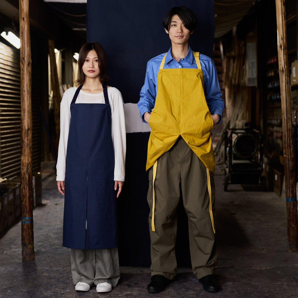 A woman in a navy Japanese apron and a man in a yellow Japanese apron stand side by side in a workshop-like setting with wooden walls.