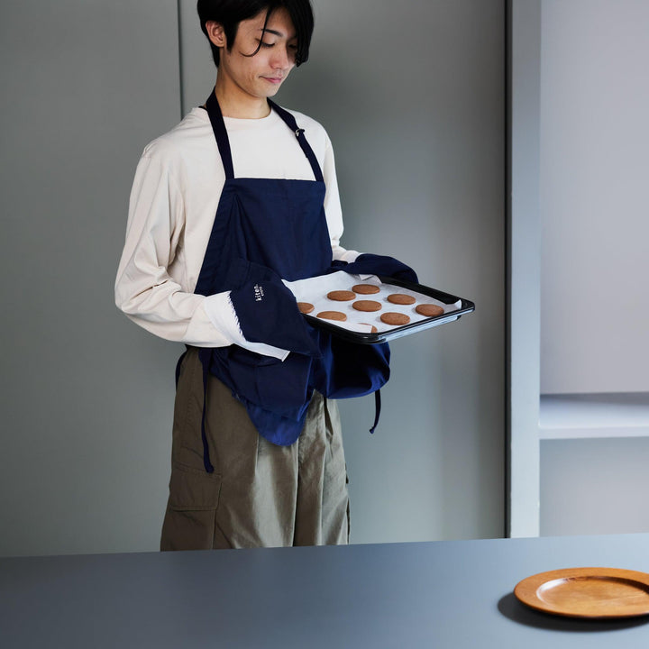 A man in a navy apron uses its built-in mitten feature to hold a tray of cookies, standing in a modern kitchen.