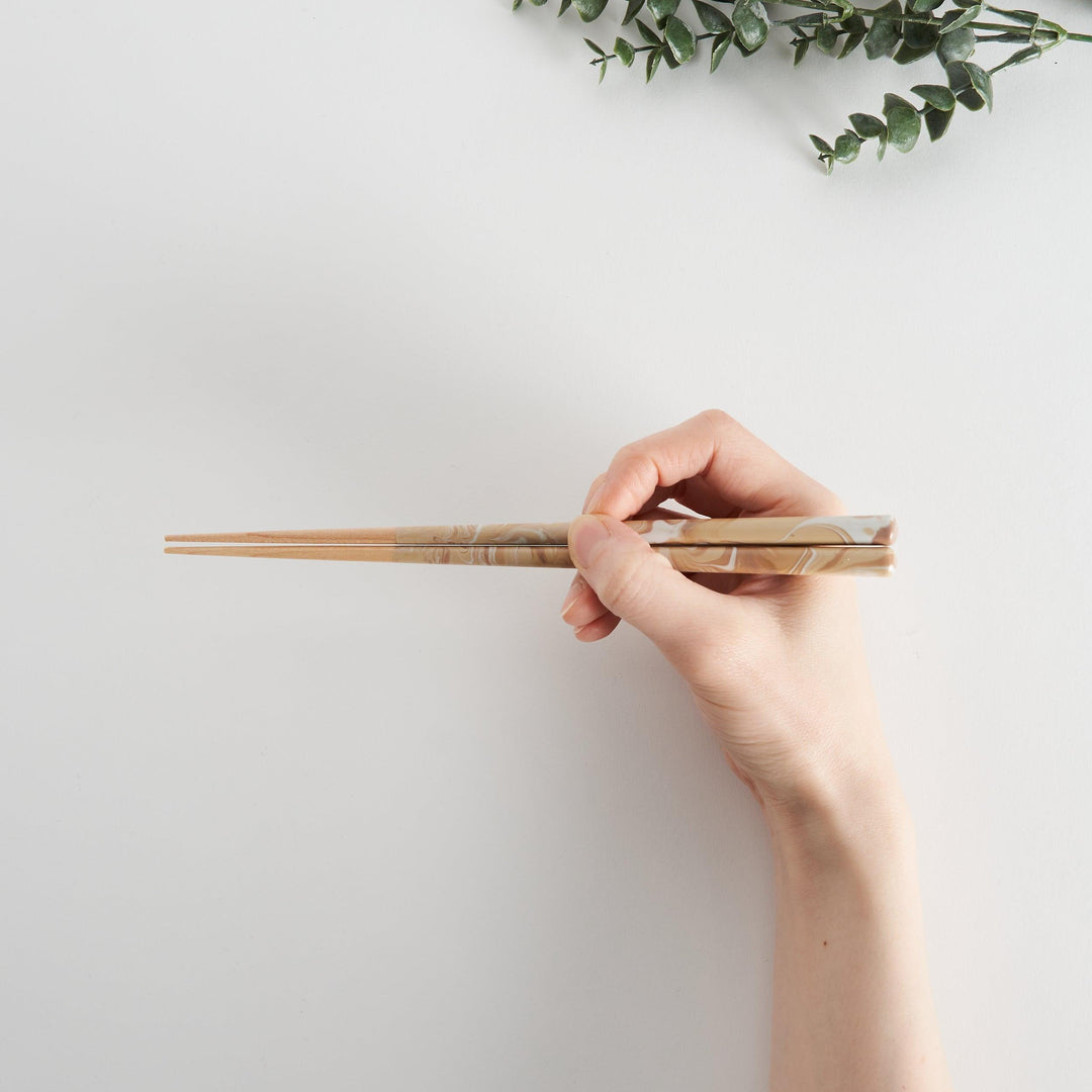 Hand holding caramel-patterned chopsticks against a light background, with eucalyptus leaves in the corner.