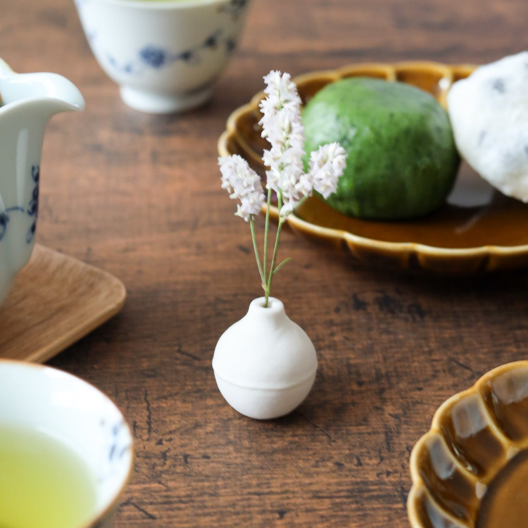 White ceramic bud vase with a round form holding tiny pale flowers, placed beside amber dessert plates and floral teacups during teatime.