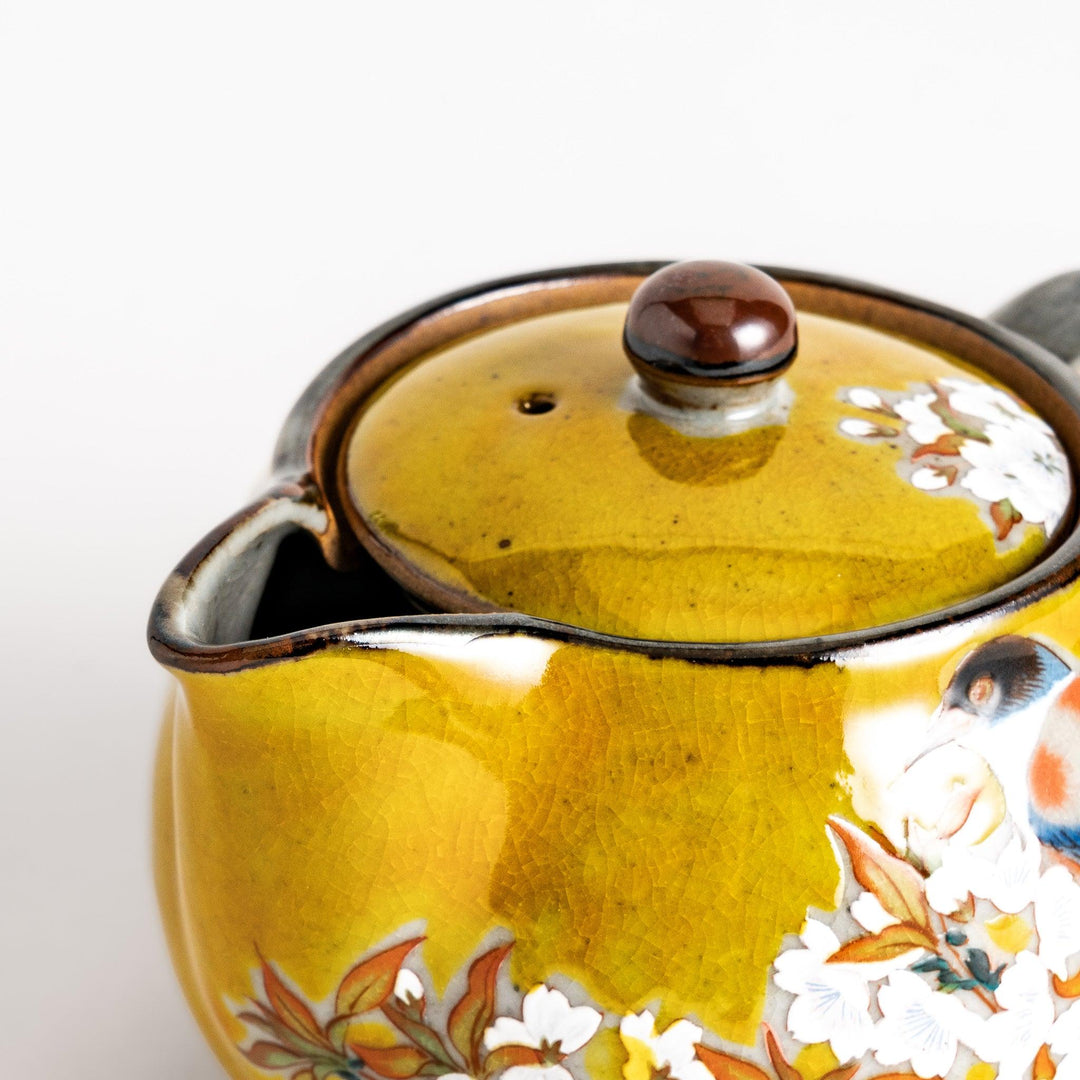 Yellow ceramic teapot with a beautiful bird and flower design, featuring a smooth sheen and secure lid, perfect for serving tea.