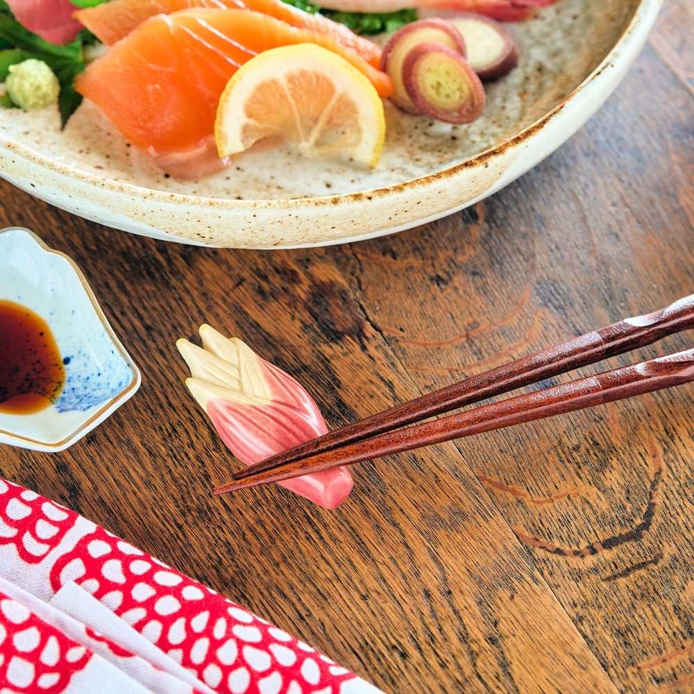 Myoga ginger-shaped ceramic chopstick rest in soft pink and cream hues, supporting wooden chopsticks next to sashimi and a blue-accented sauce dish.