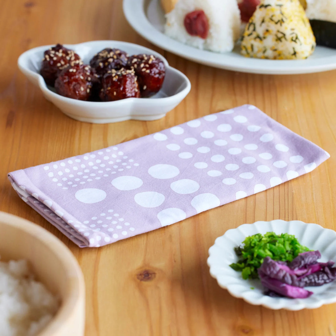 Japanese polka dot tenugui hand towel in soft purple cotton displayed on a wooden table with rice pickles and onigiri meal items